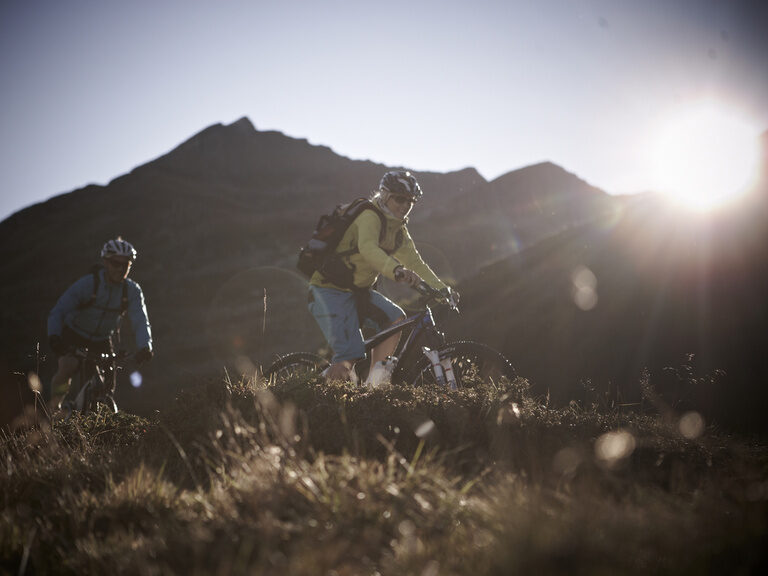 Im Vordergrund radeln zwei sportlich gekleidete Fahrer mit modernem Fahrradequipment einen Berg bei Montafon hinunter, während im Hintergrund die Sonne scheint..
