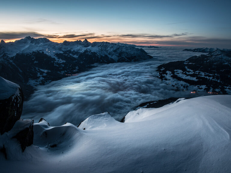 Hinter verschneiten Berggipfeln geht die Sonne unter während sich im Tal Nebel bildet.