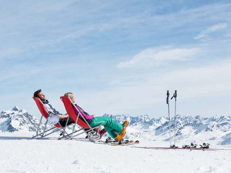 Zwei Ski-Fahrer sitzen in roten Klappstühlen und blicken über die anliegenden Berggipfel.